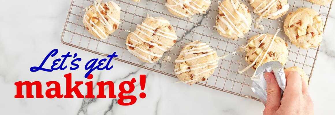 A hand decorating cooked muffin tops with icing on top of a baking rack.  - Let's get making!