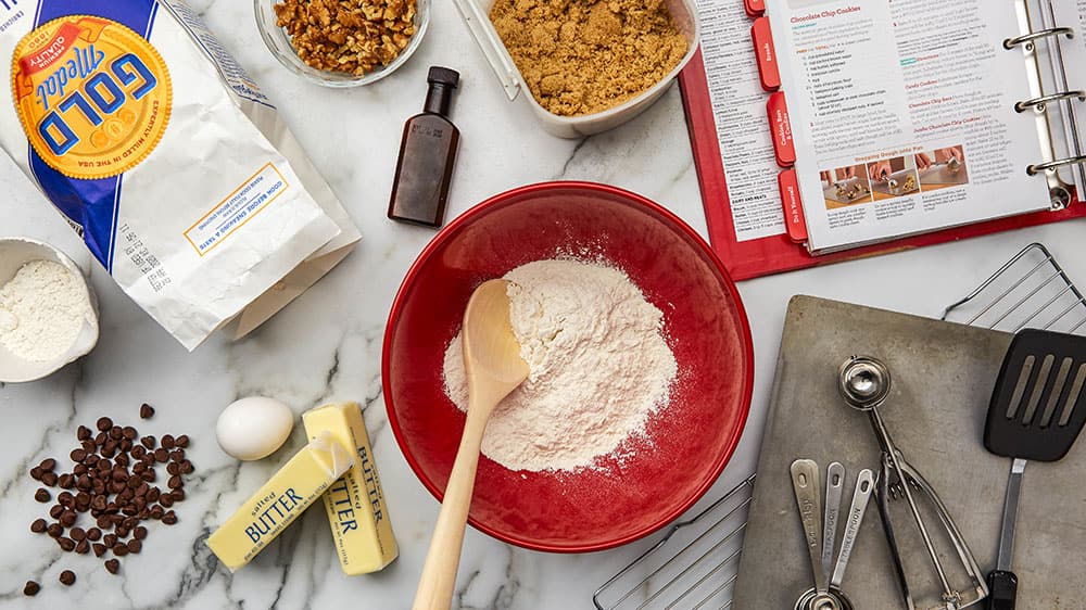 A variety of baking essentials including flour, butter, eggs, spoons, bowls